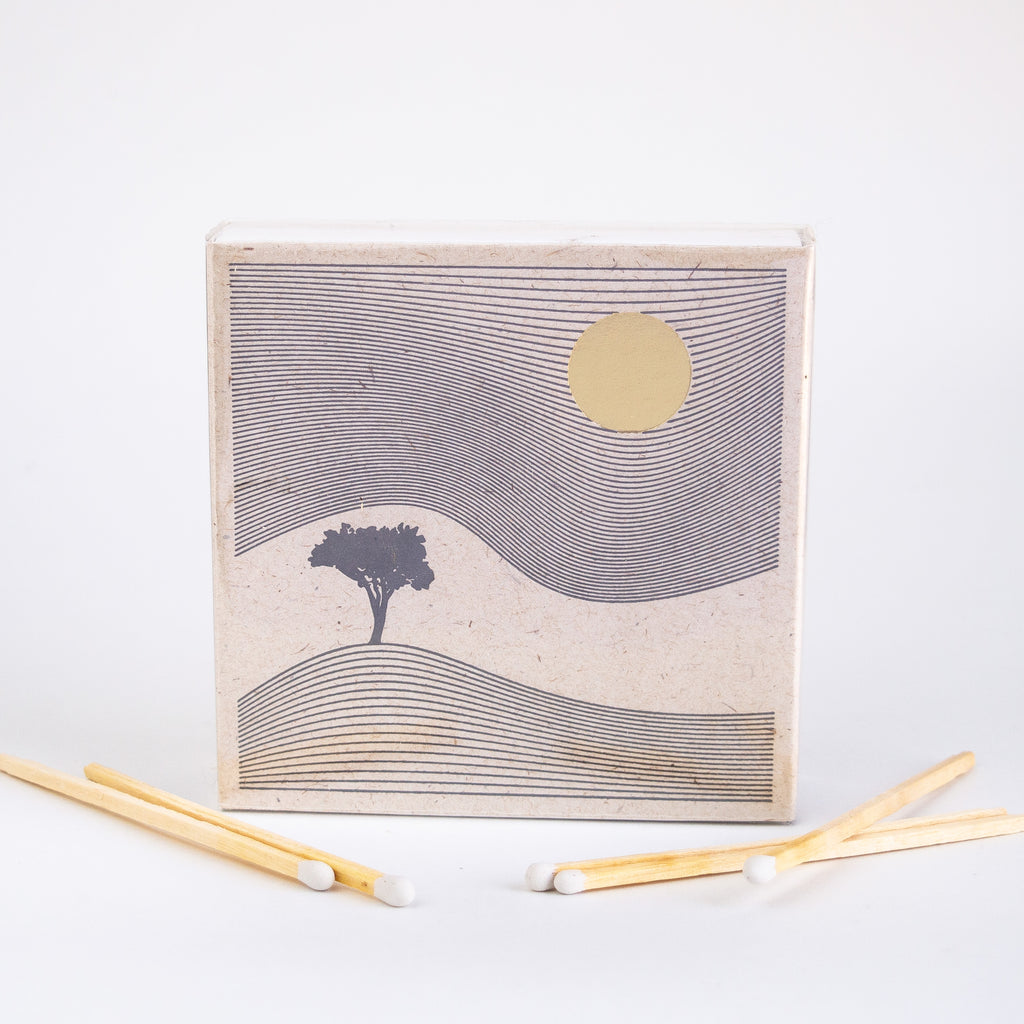 Square paper match box with a single tree sandwiched between wavy lines of land and sky, a gold sun sits in the right corner. there are various matches laying in front of the box.