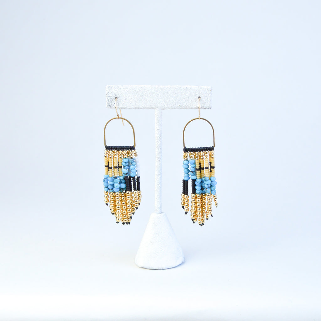 beaded fringe earrings with gold, white, black and turquoise beads in an asymmetric pattern. the beads are connected to the finding by a golden semicircle.
