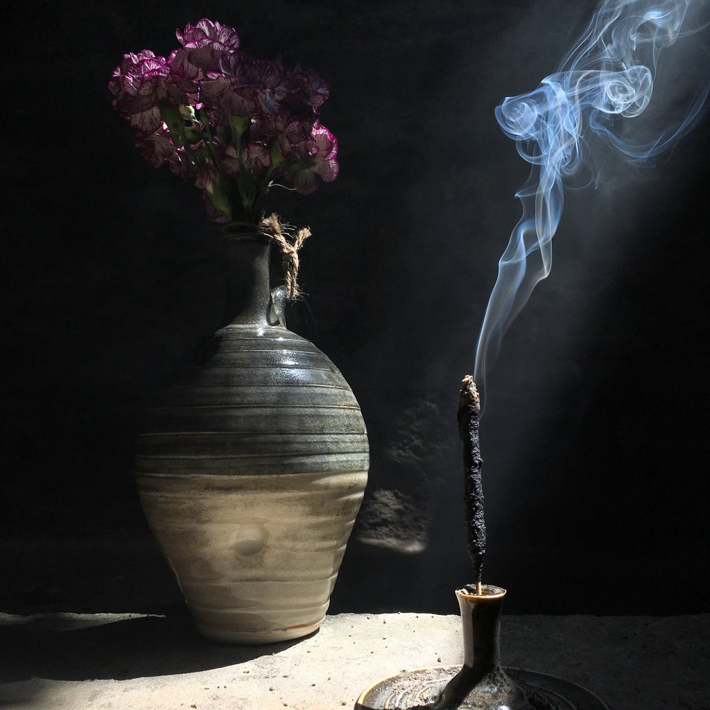 A thick palo santo incense stick lit up by a singular ray of light. Behind the incense stick is a grey pot filled with purple and white flowers.