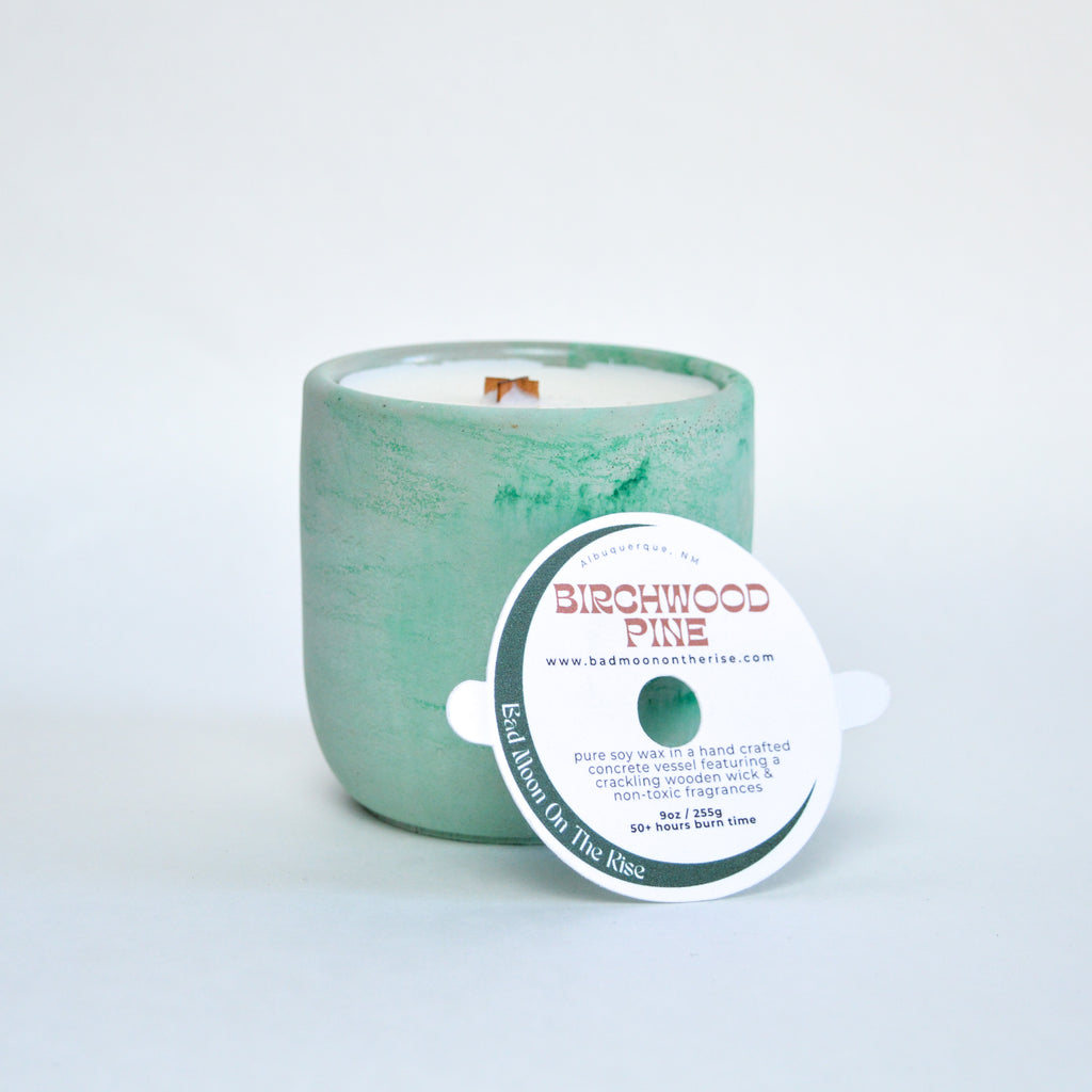 A candle with a marbled green vessel, white wax, and a wooden wick. There is a dust cover leaning against the candle with lettering that reads "Birchwood Pine"