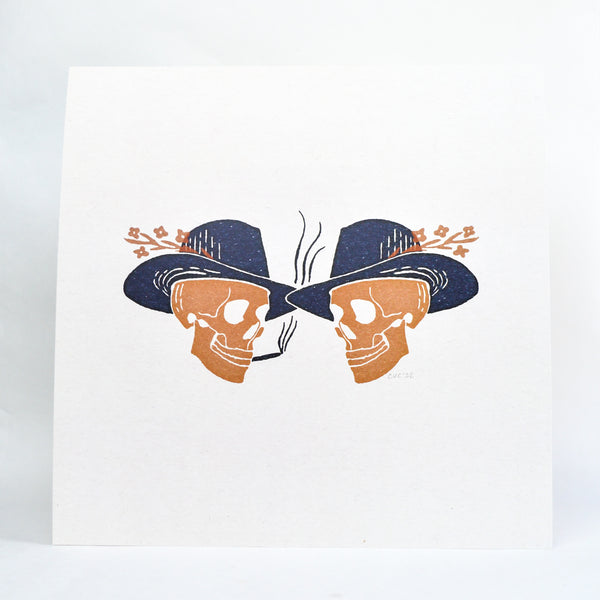 An art print with two skulls wearing cowboy hats and flowers attached to the hats. The skulls are facing each other and smiling. One is smoking.