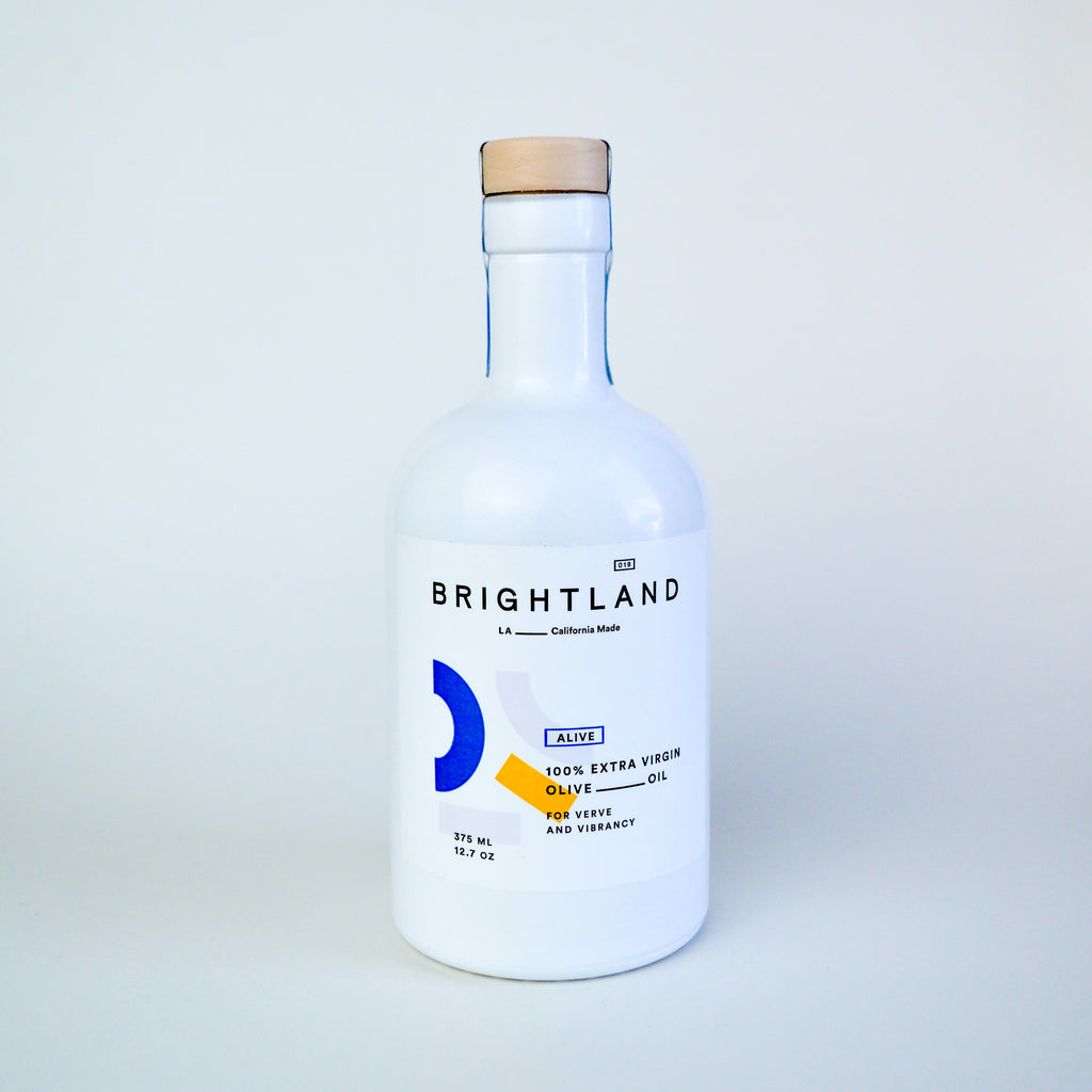 White glass olive oil bottle with a cork top on a white backdrop. There are curvy geometric shapes in the colors blue, yellow, and pink. There is text in various places around the shapes reading "Brightland LA California made" "100% Extra virgin olive oil for verve and vibrancy."