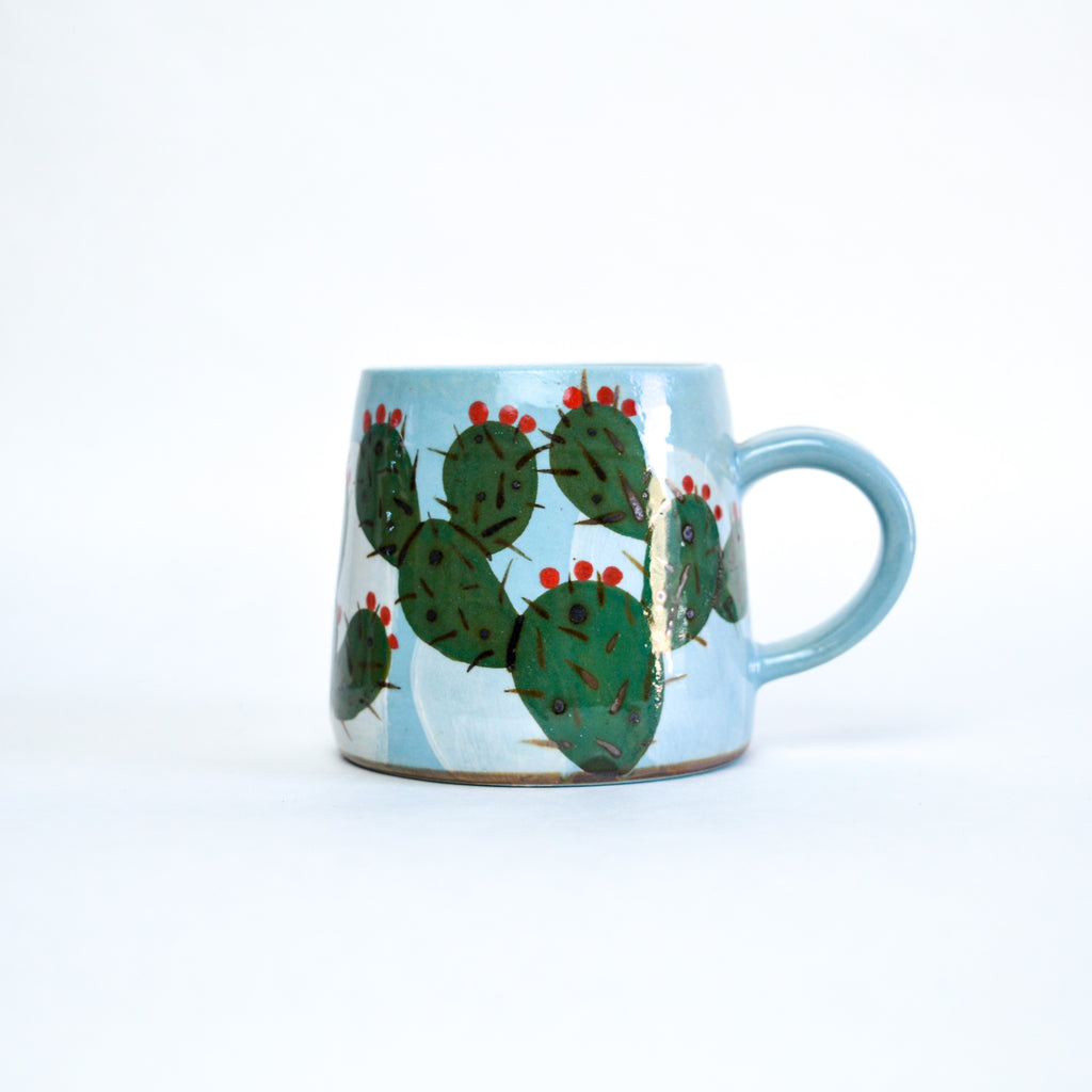 Small blue mug with a blue base. On top of the blue base are dark green cactus with black spines and red fruits.