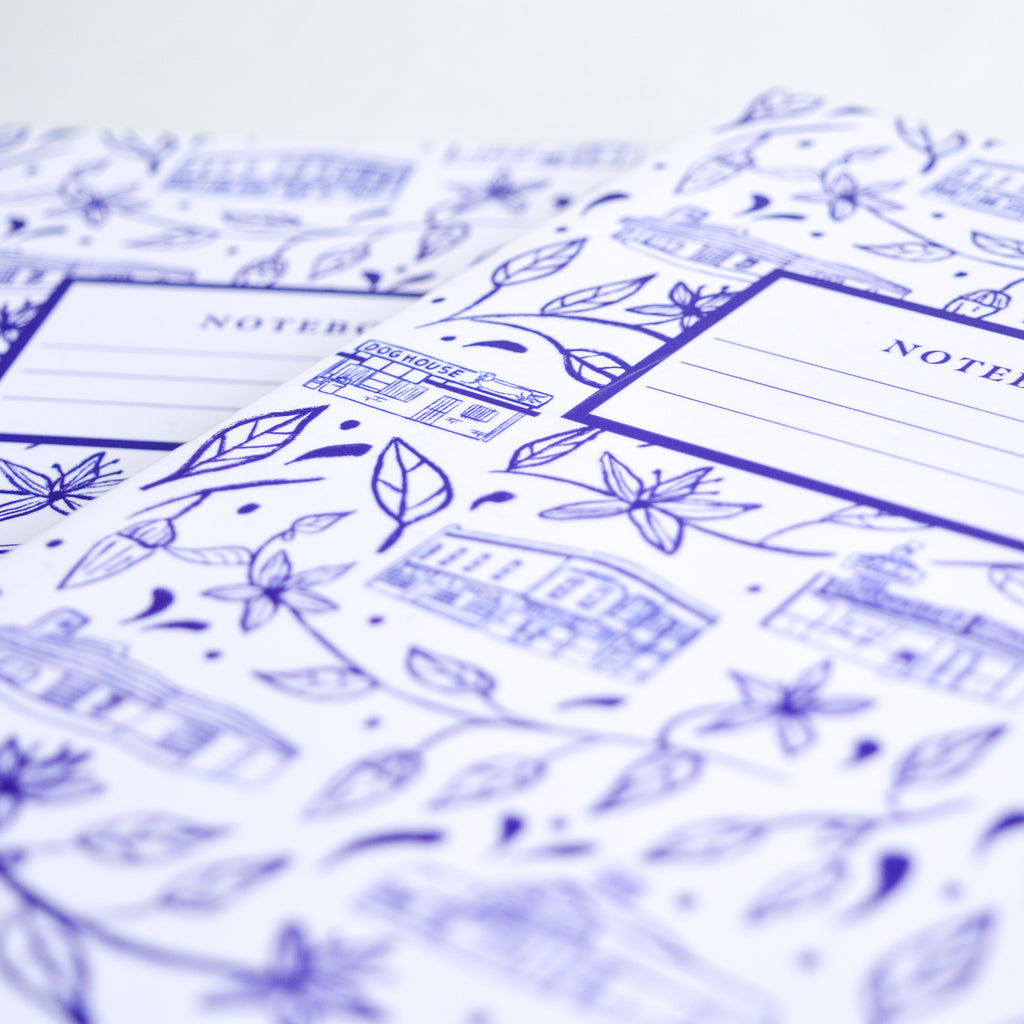 Close up of two notebooks that are fanned out. Only the covers are visible. The covers have purple art on the front of famous buildings in Albuquerque and are separated by plants and flowers. There is a white rectangle in the middle with the lettering "notebook" and three lines beneath the lettering.
