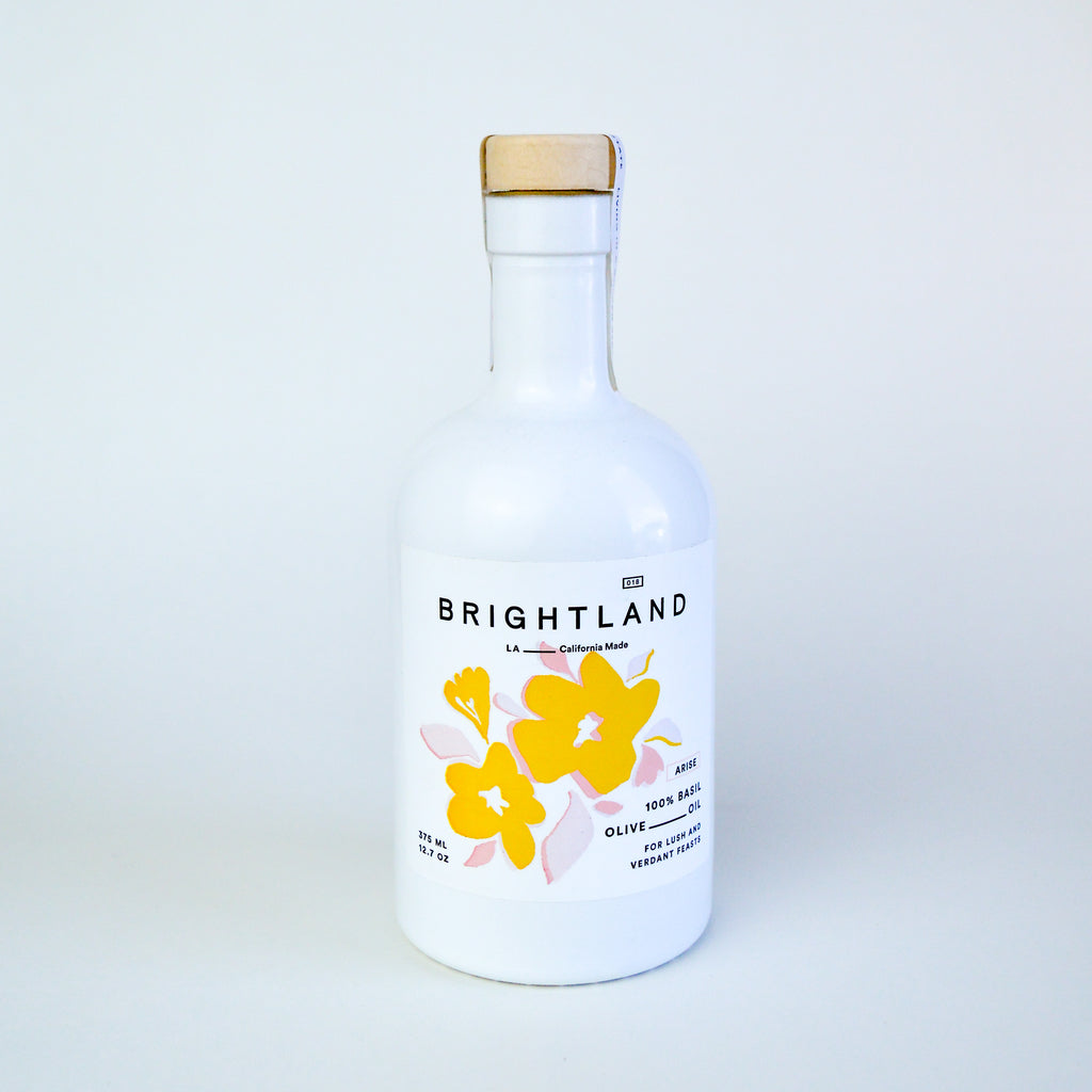 White glass olive oil bottle with a cork top on a white backdrop. There are yellow flowers with pink leaves in the middle of the label. There is text in various places around the flowers reading "Brightland LA California made" "100% basil olive oil for lush and verdant feasts."