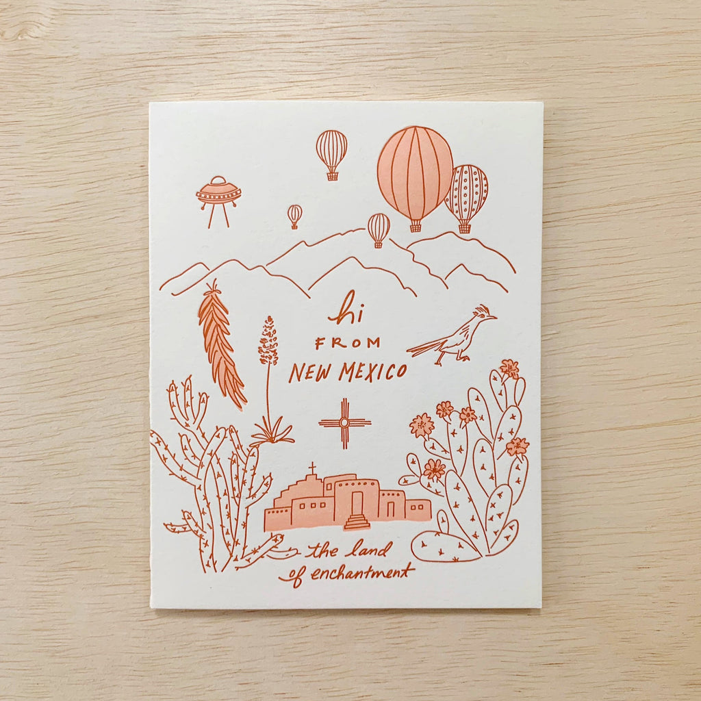 White card with orange mountains, hot air balloons, chiles, a road runner, cactus, and adobe church. There's orange writing which reads "Hi from New Mexico, the land of enchantment."