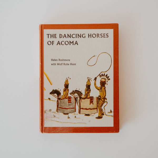 A cover of the book "The Dancing Horses of Acoma" by Helen Rushmore and Wolf Robe Hunt. The cover is a drawing of three people from the Pueblo of Acoma. Two on horses and one carrying a  whip, wearing a large feathered headdress. 