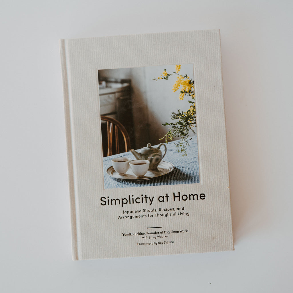 A cover of the book "Simplicity at Home" by Yumiko Sekine. The cover is white with a photograph of a tea set and a flower arrangement on a kitchen table. Additional text reads "Japanese Rituals, Recipes, and Arrangements for Thoughtful Living."