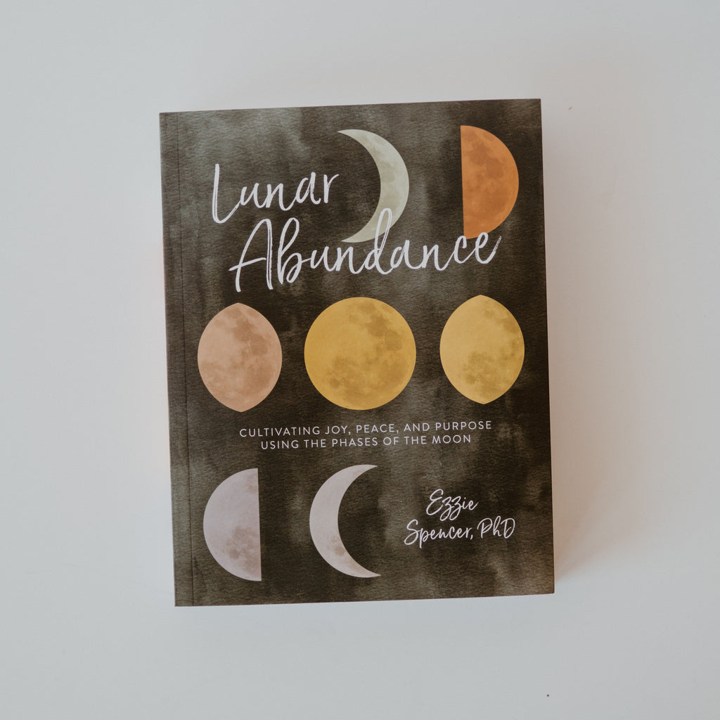 A cover of the book "Lunar Abundance" by Ezzie Spencer. The cover is black with different colored illustrations of phases of the moon. Additional text reads "Cultivating Joy, Peace, and Purpose Using the Phases of The Moon."