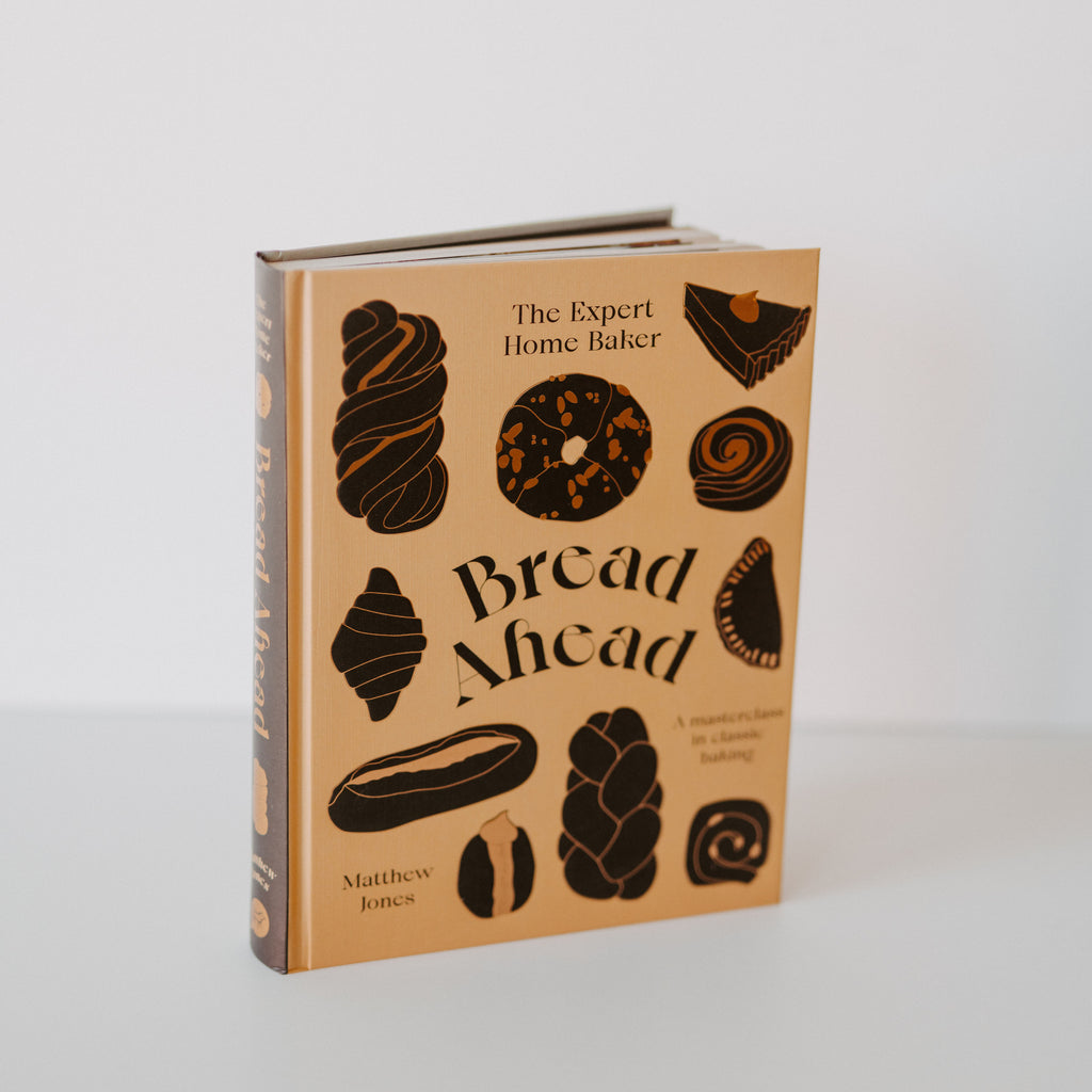 The book "Bread Ahead" by Matthew Jones standing on a white backdrop. The has a tan base color and various pastries in a dark brown color. There is other text on the book reading "the expert home baker" "a masterclass in classic baking."