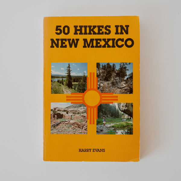 Yellow Book reading "50 Hikes in New Mexico" by Harry Evans. Below the title are four pictures in a quadrant formation of varying landscapes in New Mexico. Directly in the middle of the landscape photos is an orange Zia symbol.
