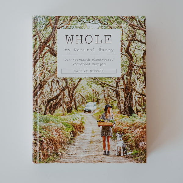 The cover of the book "Whole" by Harriet Birrell. The cover is an image of a woman standing amongst a tree lined dirt road next to a dog, while holding a crate of plants. Text reads "Whole by Natural Harry: Down-to-earth plant-based wholefood recipes."