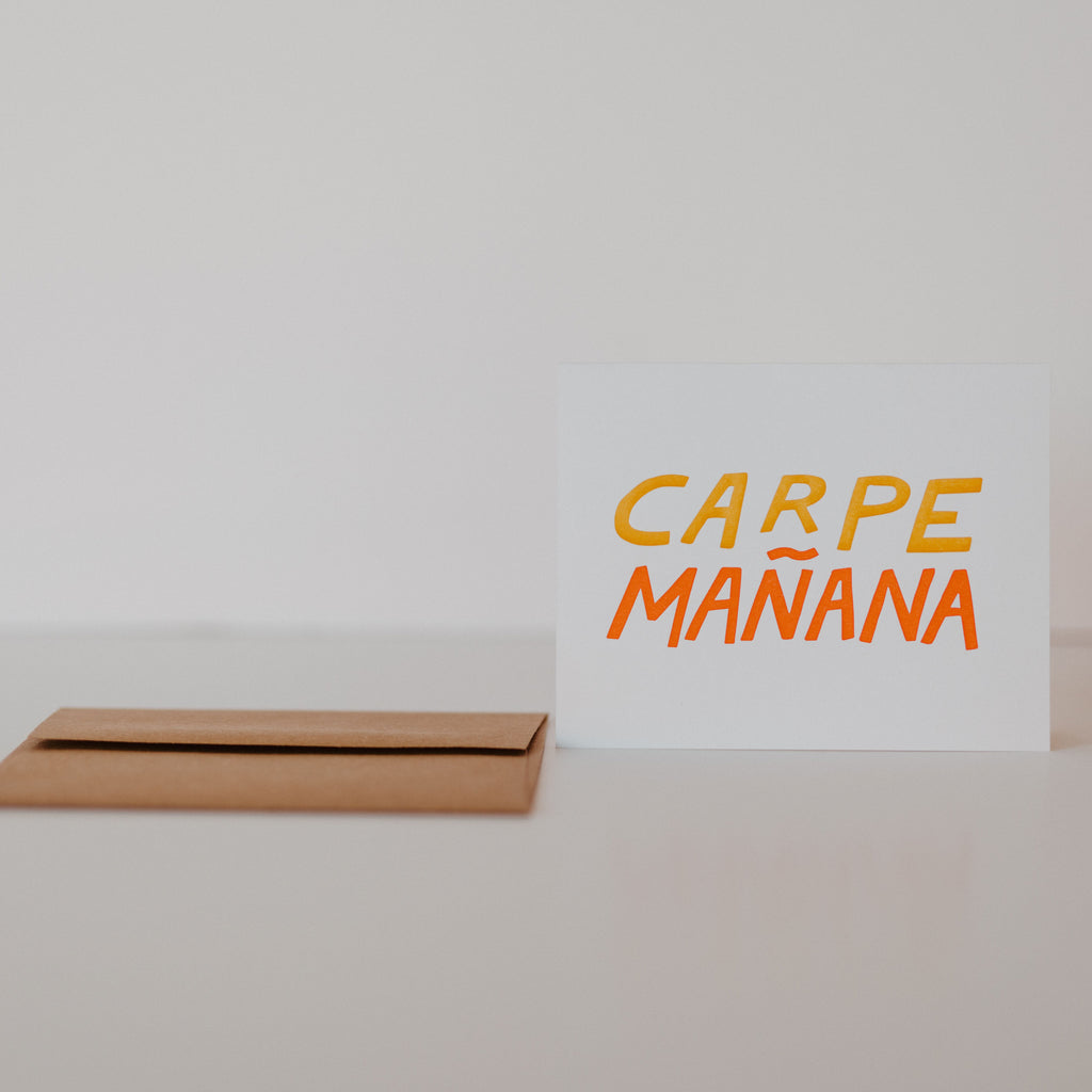 Dark beige envelope laying on a white backdrop with a white card standing next to it. The card has yellow and orange lettering in the middle reading "carpe mañana."