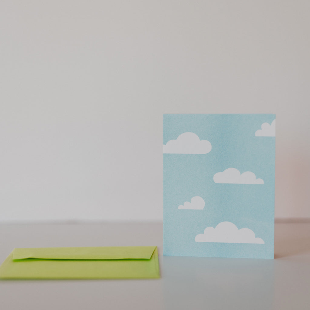 Green envelope laying on a white backdrop with a blue card standing next to it. There are  white cartoon clouds on the blue card.