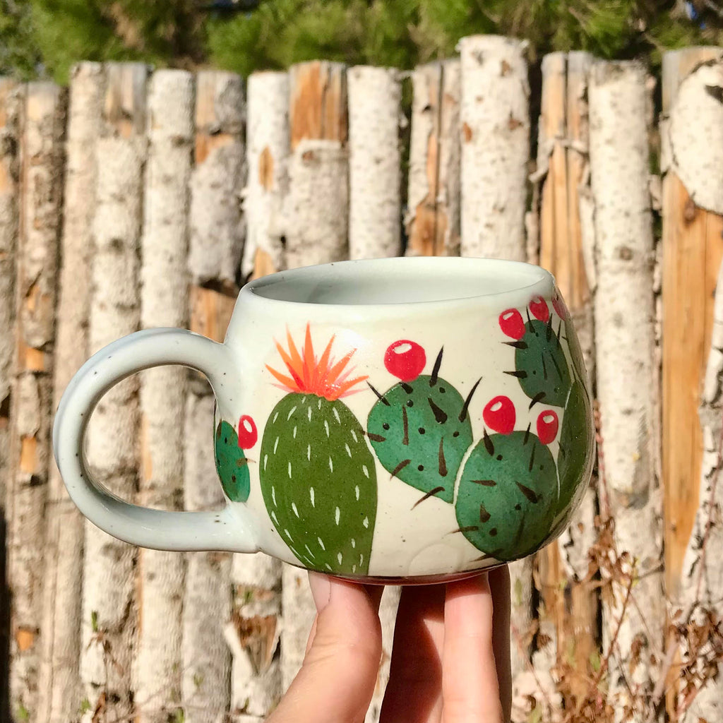 Very small, round mug with a white base. On top of the base are cacti with varying shades of green. Most with prickly pear and one with an orange flower.