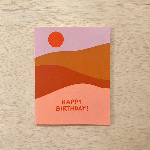 Happy Birthday card with different layers of colors to look like sand dunes. Purple Sky, camel, rust red and light sand pink layered colors. An orange red son is printed in the sky. It says Happy Birthday! 