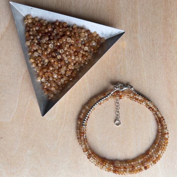 A wrap bracelet and triangle tray full of beads on a wooden background. The triangle tray (in the rightmost corner) is steel and filled with the same beads the wrap bracelet is made out of. The bracelet is in the leftmost part of the photo. The bracelet has a variation of tan beads and has a silver closure.