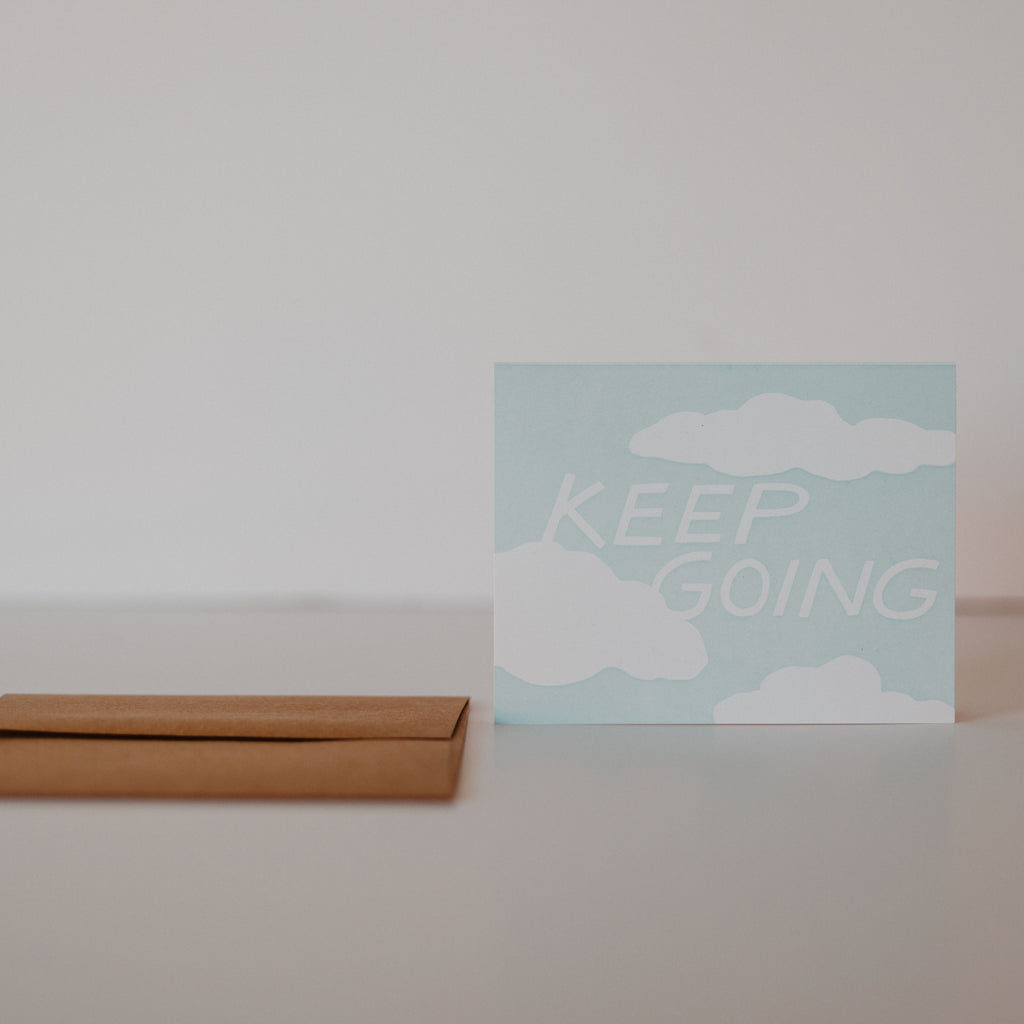 Dark brown envelope laying on a white background with a light blue card standing next to it. The blue card has white clouds and white text in the middle reading "Keep going."