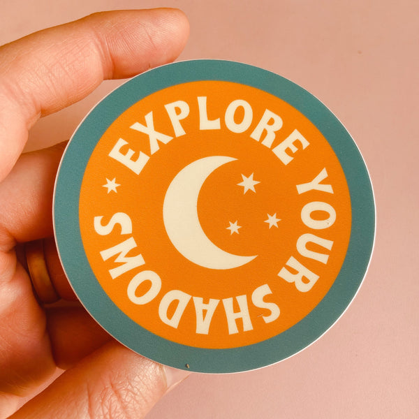 Circular sticker with a blue boarder and orange middle. there is a white moon in the middle and white text around it reading "explore your shadows."
