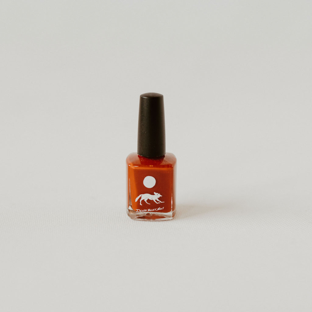 Burnt orange nail polish with a black top. In the middle of the nail polish bottle there is a moon with a coyote under it and text that reads "Death Valley Nails" all are in white.