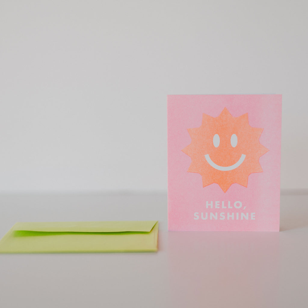 Pink card with lime green envelope. the card has a salmon colored sun with a white happy face and white text under it reading "Hello Sunshine"