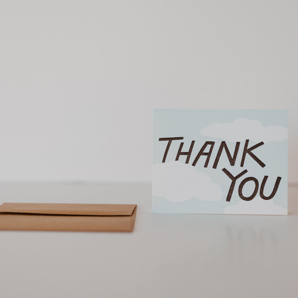 Brown envelope laying on a white background with a blue card standing next to it.  The card has white clouds and black text reading "thank you"