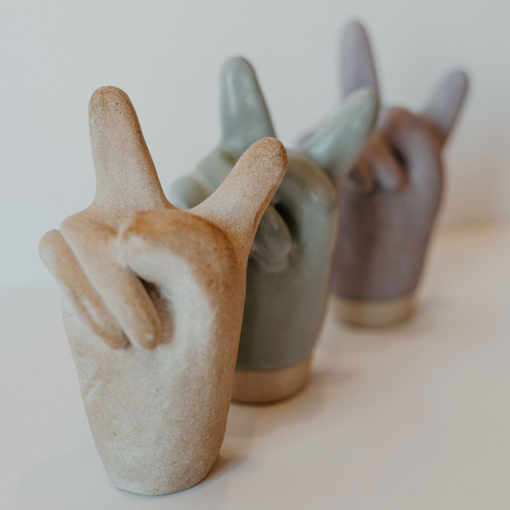 3 ceramic peace sign hands in beige, blue, and purple. Each color is a separate hand standing up where the wrist would be.