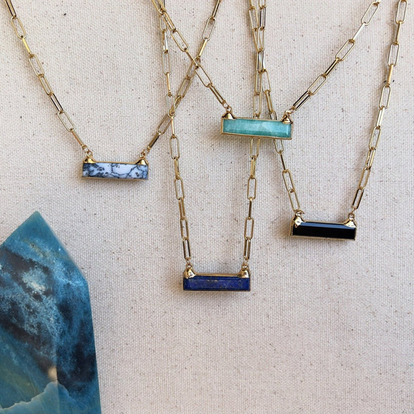 Four Gold necklaces with a paperclip style chain. There are semi precious stones in a rectangular shape horizontally connected to each necklace. From left to right: Amazonite, Lapis Lazuli, Black Onyx, and Dendretic Agate