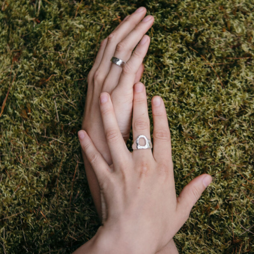 someone wearing a silver, rough hammered ring with an empty circle in the middle of it. The persons hands are laying organically on small green leaves.