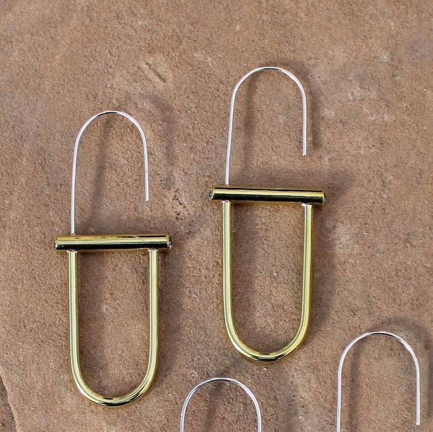 Brass earrings laying next to one another. the earrings have an oval shape with a barbell in the middle and a thin open loop at the top.