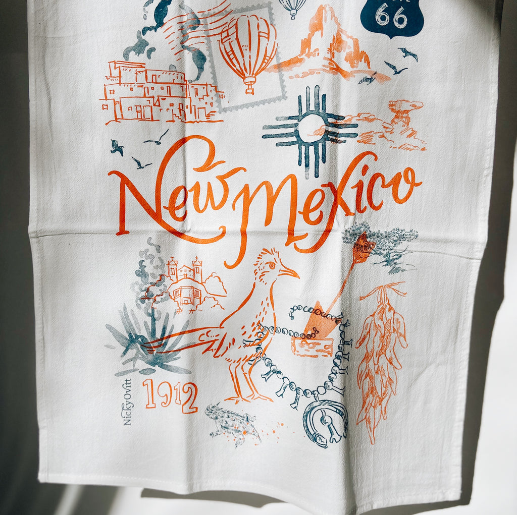White tea towel with southwestern animals and landmarks in multiple colors placed about the towel. There's red text in the middle reading "New Mexico."