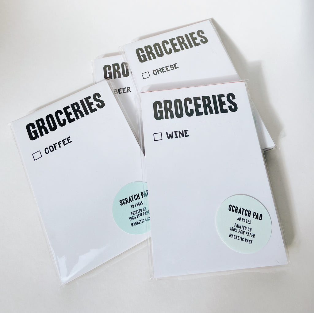 Multiple white scratch pads with black lettering reading "groceries." Under that is a checkbox with one food item written down. from top to bottom: wine, coffee, cheese, beer.