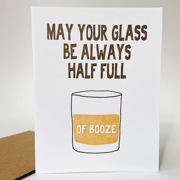 A white card with brown text reading "may your glass always be half full"  and white text reading "of booze." the white text is in a doodled scotch glass filled with amber liquid.