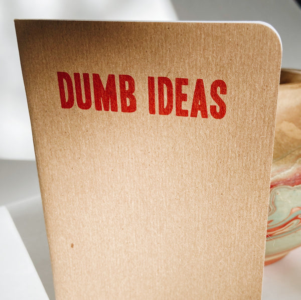Brown paperback notebook with red lettering at the top reading "dumb ideas."