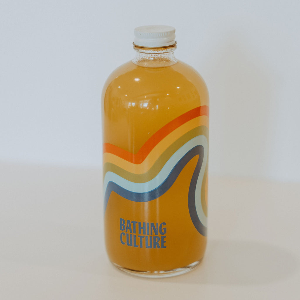 Glass bottle with a white metal cap and yellow soap inside. The bottle has a pastel rainbow and blue text reading "Bathing Culture"