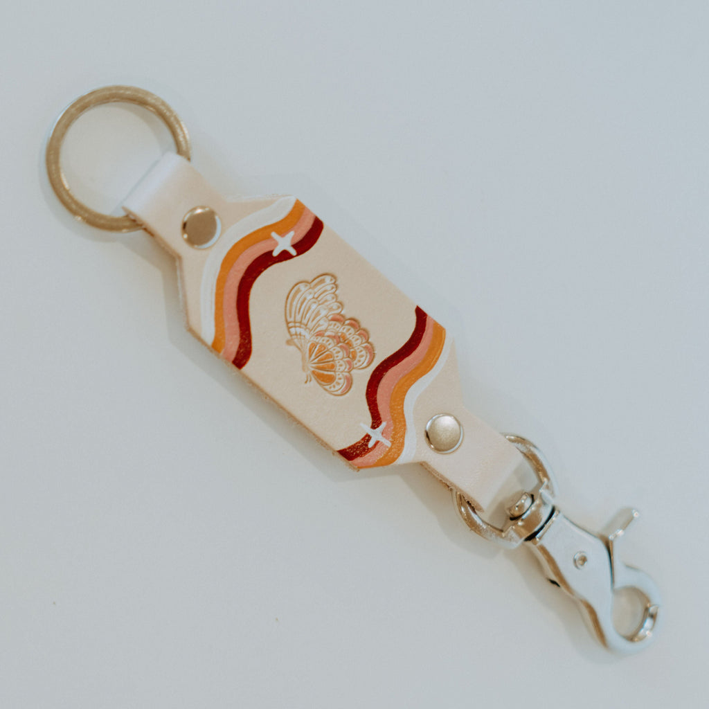 Pink leather keychain with a silver key ring on top and a silver snap hook on the bottom. There is a butterfly stamped in the middle of the leather which is painted white and orange. Four curvy lines of white, orange, pink, and maroon painted on either end with a white star on top of them.