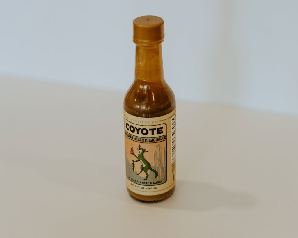 Glass hot sauce bottle with a beige label. the label has a green coyote holding fire in one paw and chiles in the other. theres black writing reading "Coyote, roasted green magic sauce. la salsa verde magica."