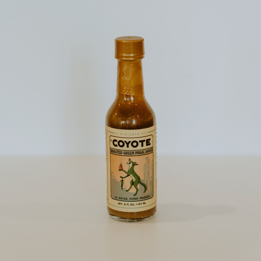 Glass hot sauce bottle with a beige label. the label has a green coyote holding fire in one paw and chiles in the other. theres black writing reading "Coyote, roasted green magic sauce. la salsa verde magica."