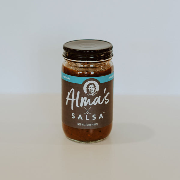 Glass jar of salsa with black aluminum lid. There is a label running horizontally across the middle of the jar. at the top of the label is a light blue strip with the lettering "medium" in white. Below that on a brown background is a picture of a middle aged woman. Below, in white lettering, the rest of the label reads "Alma's Salsa" "net WT. 16 oz (454G)."