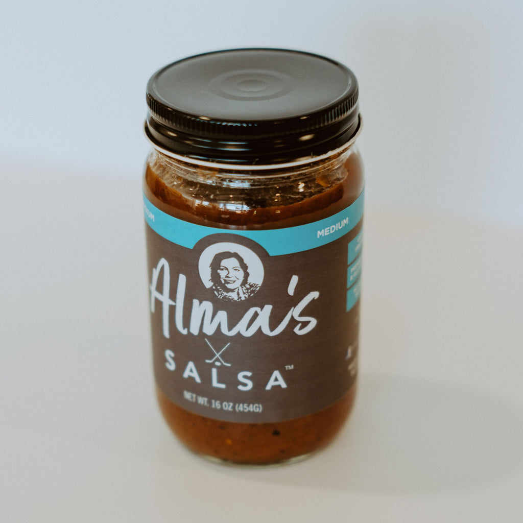 Glass jar of salsa with black aluminum lid. There is a label running horizontally across the middle of the jar. at the top of the label is a light blue strip with the lettering "medium" in white. Below that on a brown background is a picture of a middle aged woman. Below, in white lettering, the rest of the label reads "Alma's Salsa" "net WT. 16 oz (454G)."