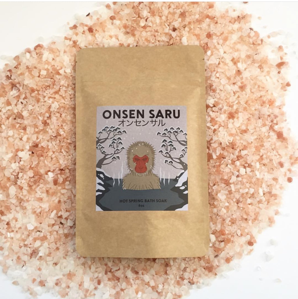 Brown paper bag with a blue label and black text reading "Onsen Saru hot spring bath soak." The bag is laying atop a pile of pink bath salt.